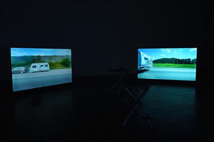 Zu sehen ist ein dunkler bzw. durch zwei Monitore schwach belichteter Raum. Auf den Monitoren sind jeweils ein Auto/Camper auf einer Autobahn mit grüner Landschaft zu sehen. 